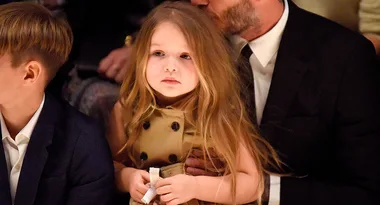 A young girl with long hair sits on an adult's lap at an event, surrounded by other people.