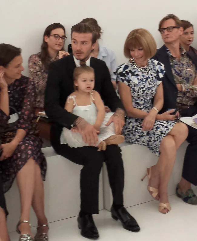 Man in a suit with a child on lap seated next to a woman in a patterned dress at a fashion event.