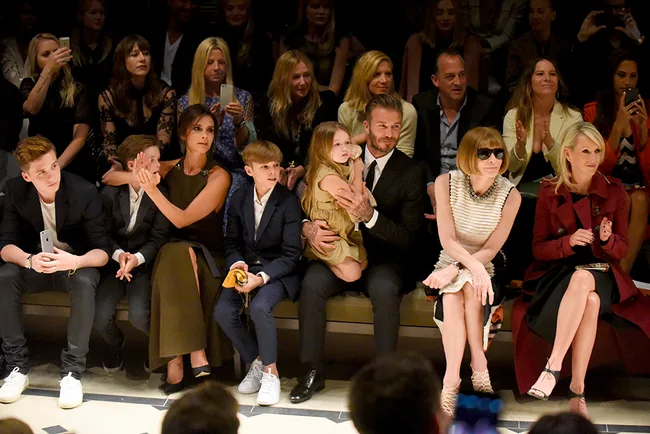 A group of people, including a family and others, sit in the front row at a fashion show, engaged with the event.