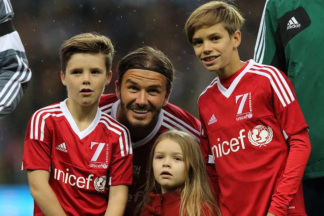 Former footballer with long hair poses with three children in red UNICEF jerseys on soccer field.