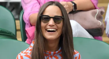 A woman with long brown hair and sunglasses smiles while seated in a green chair.