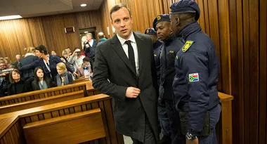 Man in a suit escorted by police in a courtroom while people watch from the gallery.