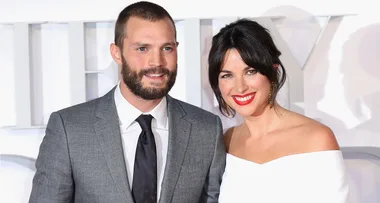 A smiling couple poses on the red carpet; the man wears a suit, and the woman wears a white off-shoulder dress.
