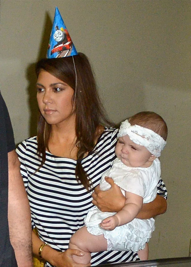 Woman wearing a striped top and party hat holds a baby with a headband, both looking in the same direction.