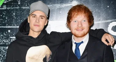 Two men standing together, smiling; one in a gray cap and hoodie, the other in a suit with a blue tie.
