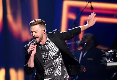 A man singing passionately on stage with one hand raised, wearing a patterned shirt and blazer.