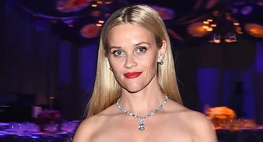 ```plaintext
Person with long blonde hair, wearing a diamond necklace, at a formal event with a dark, colorful background.
```