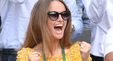 A woman in sunglasses cheers excitedly, wearing a yellow dress at an outdoor event.