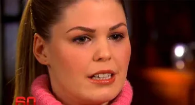 A person with long hair wearing a pink turtleneck, speaking during an interview on TV.