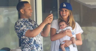 A man takes a photo with a phone while a woman holds a baby, both wearing casual clothing and smiling indoors.