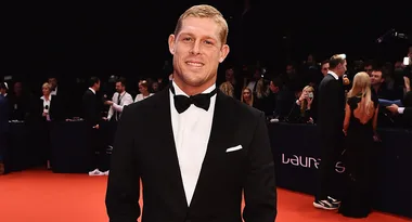 Man in tuxedo smiling on red carpet at an event with crowd in background.