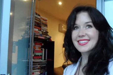 Young woman with dark hair smiling indoors near a stack of books.