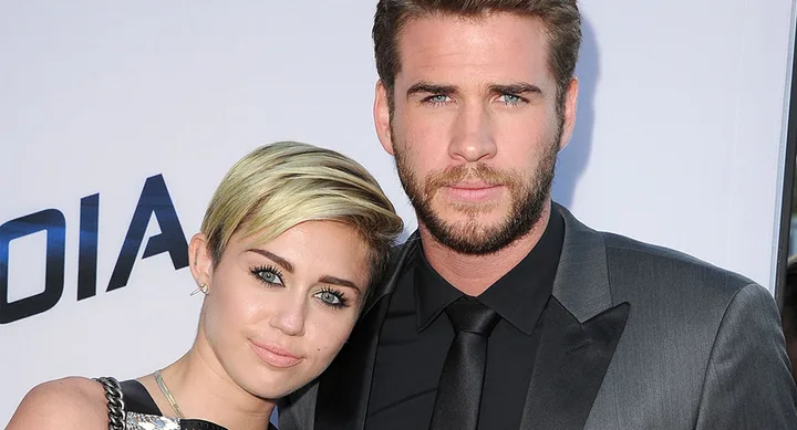 Two people posing together, one with short blonde hair and the other with a beard, both dressed formally.