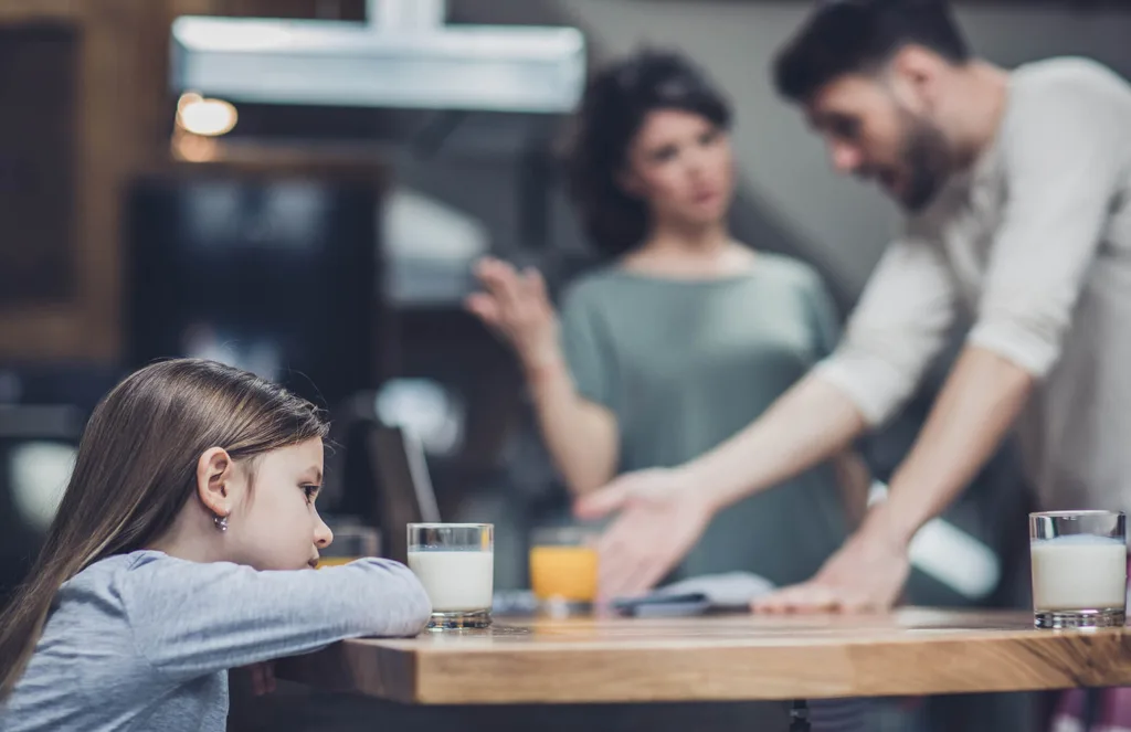 Young girl sitting on table while a couple fights in the background