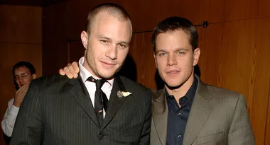 Two men in suits pose together at an event, with one man placing his arm around the other's shoulder.