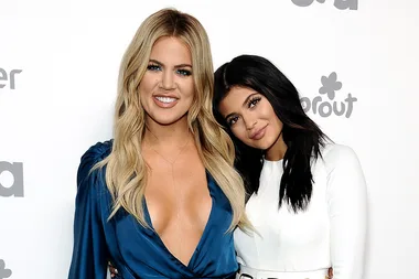 Two people pose together, smiling, on a red carpet event. One wears a blue dress, the other a white outfit.