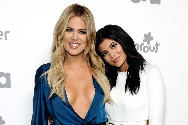 Two people pose together, smiling, on a red carpet event. One wears a blue dress, the other a white outfit.