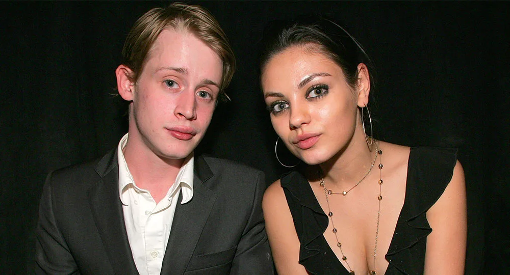 Two people posing together, the man in a suit and the woman in a black dress with hoop earrings, against a dark background.