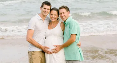 A pregnant woman in a white dress stands on the beach between two men, all smiling at the camera.