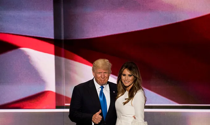 Two people posing in front of a large American flag, one giving a thumbs-up.