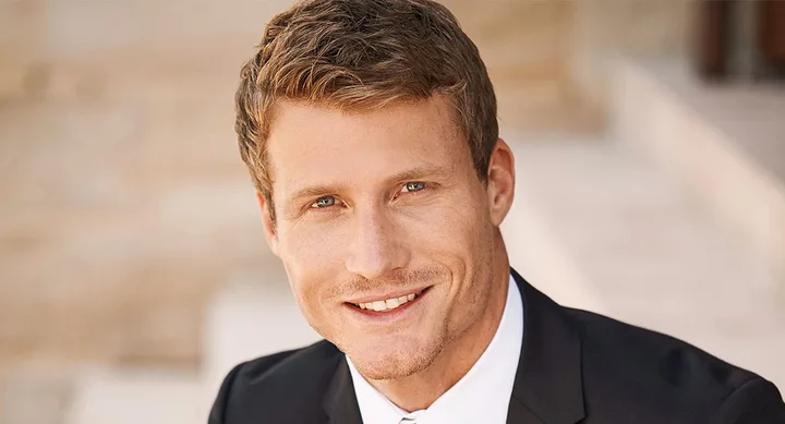 Man in a suit smiling outdoors with blurred background.