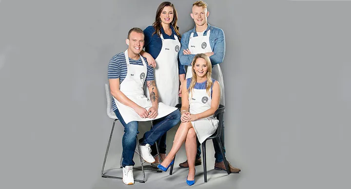 Four people wearing white aprons with a logo, posed against a gray background, two standing, two seated.
