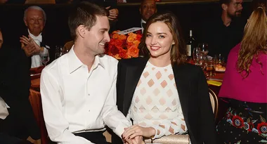 A couple smiling and holding hands at a formal event, seated at a table with flowers and drinks.