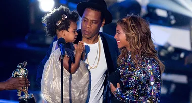 Three people on stage, one holding a child, with a trophy handed over. Wearing stylish formal attire under stage lights.