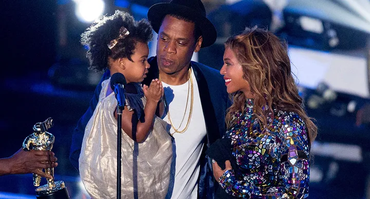 Three people on stage, one holding a child, with a trophy handed over. Wearing stylish formal attire under stage lights.