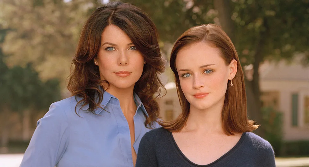 Two women standing outdoors, one with long dark hair in a blue shirt and the other with straight brown hair in a dark top.