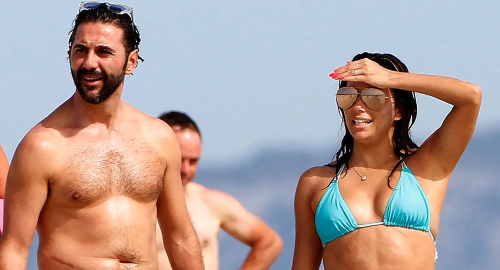 Man and woman in swimwear on a sunny beach; woman shielding her eyes with her hand, both appear relaxed.