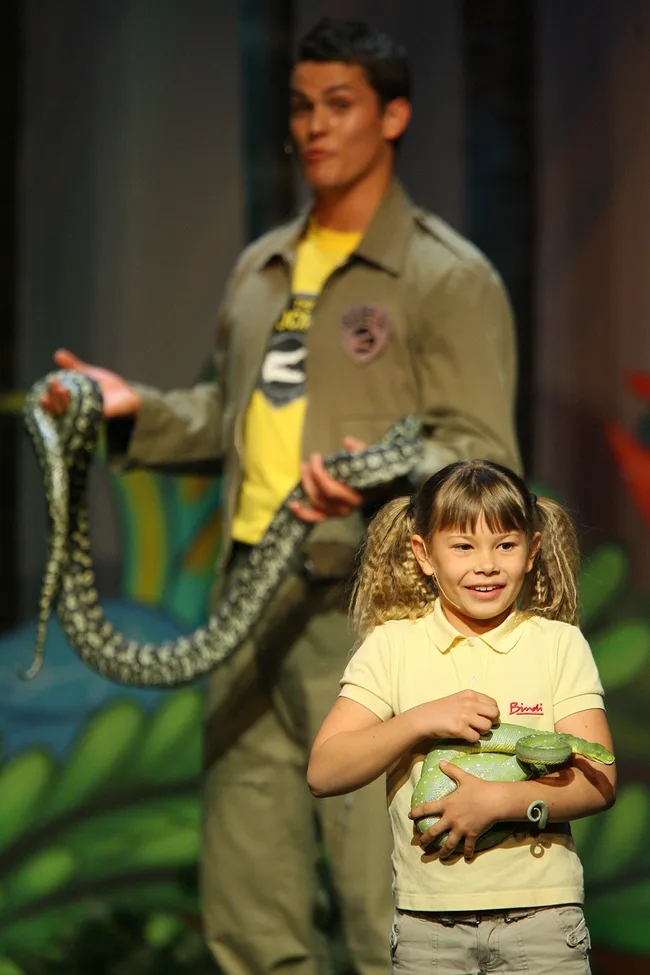 Girl holds a lizard while a person in the background holds a snake, smiling during a wildlife presentation.