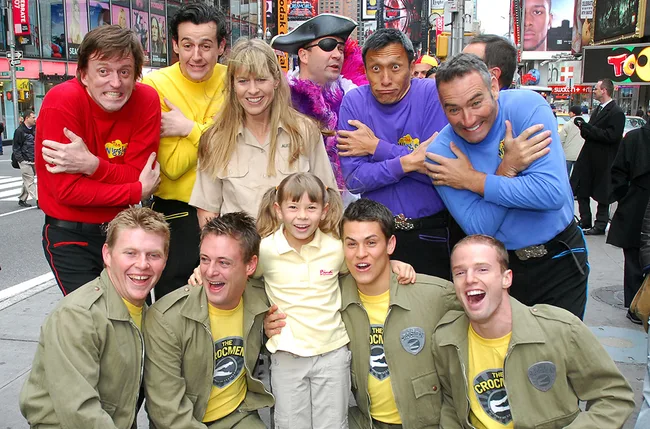 Group photo of people in colorful shirts and costumes, including men and women, smiling and posing in a busy urban setting.