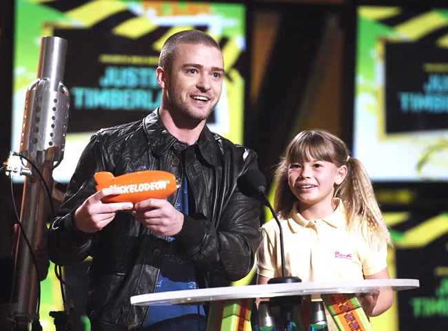 Two people on stage at a Nickelodeon awards show, one holding an orange blimp trophy.
