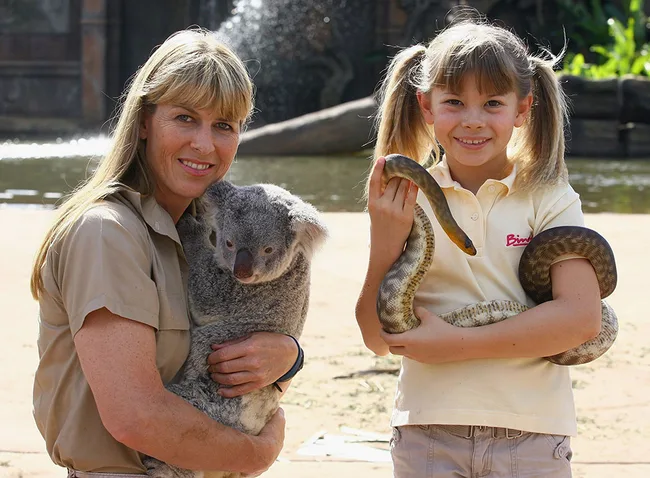 Two people smiling, one holding a koala and the other holding a snake, outdoors with a water feature in the background.