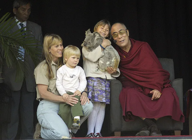A family with a koala sits with a man in a maroon robe.
