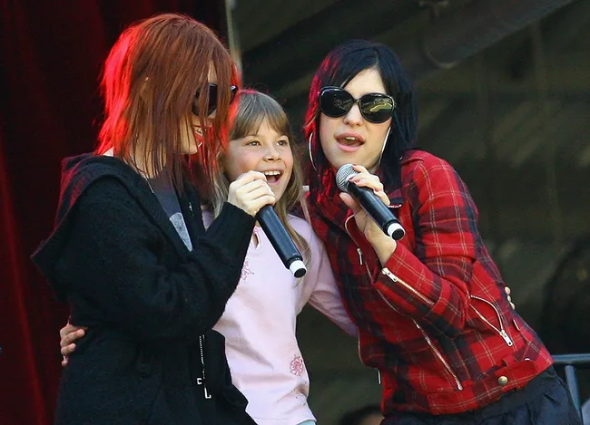 Two women performing on stage with a child, all holding microphones and smiling.