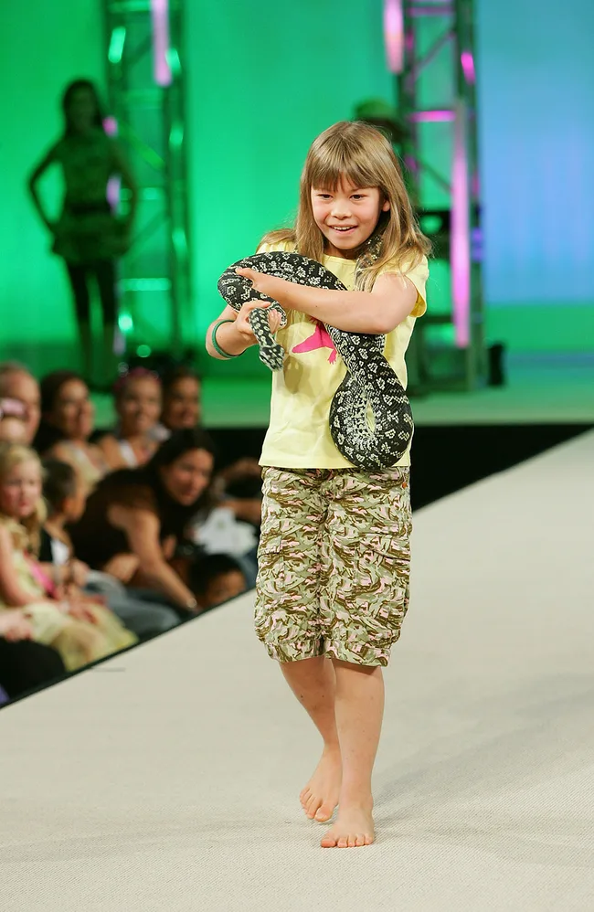 Girl in camo shorts holding a black and white snake, walking barefoot on a runway, with audience in the background.