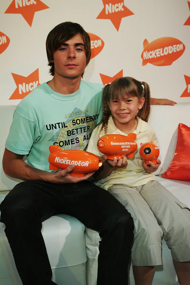 Young man and girl holding Nickelodeon awards, sitting on a white couch at a Nick-themed event.