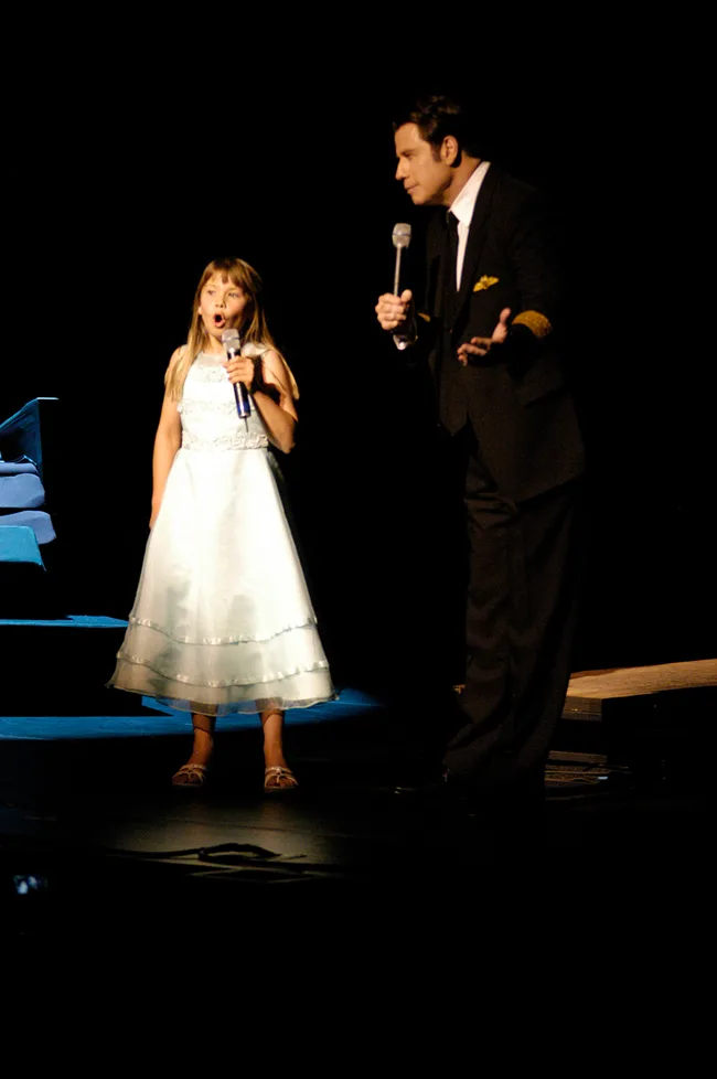 A young girl in a white dress and a man in a suit sing on stage with microphones.