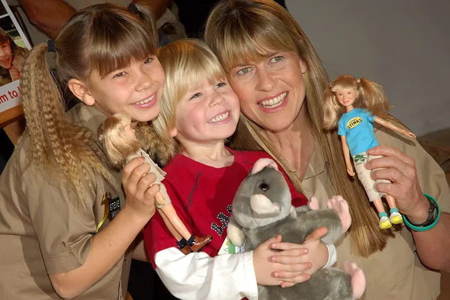 Two children and a woman smiling, holding dolls and a stuffed toy.