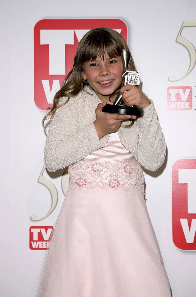Young girl holding a TV Week award, smiling, in a pink dress with floral embroidery at an event.