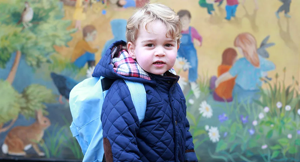 A young child in a quilted jacket with a blue backpack stands in front of a colorful mural with nature and children.