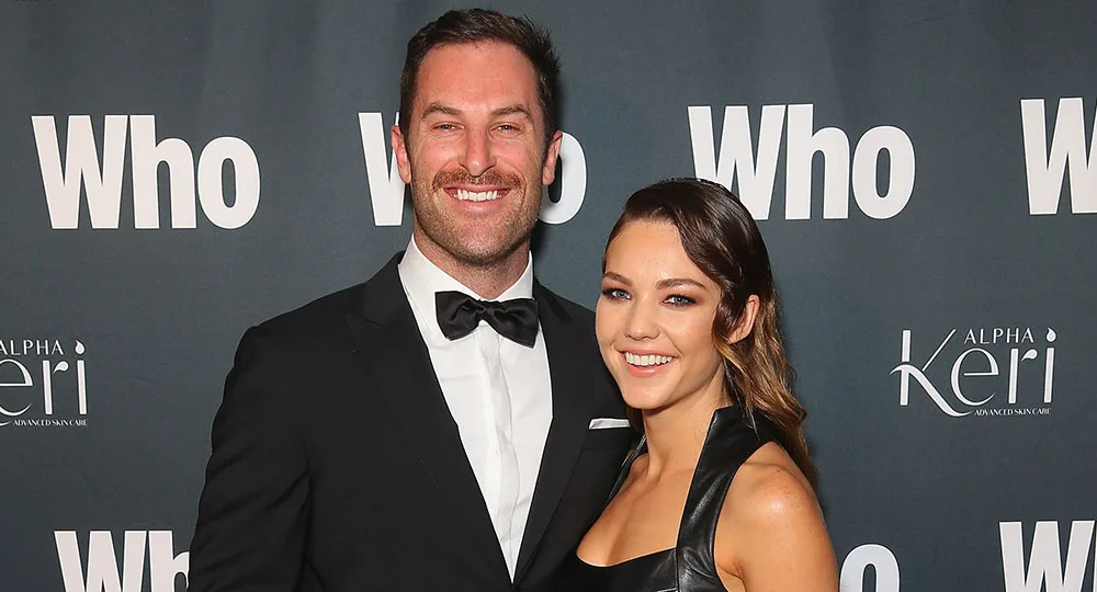 A couple on red carpet, man in black suit, woman in black dress, smiling, "Who" backdrop.