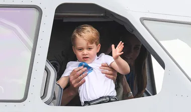 Young child waving from a helicopter cockpit, held by an adult, with a woman in the background.