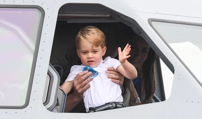 Young child waving from a helicopter cockpit, held by an adult, with a woman in the background.