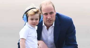 A man in a blue jacket leans close to a child wearing a white shirt and blue headphones.