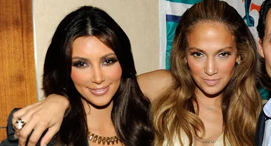 Two women smiling, one with long dark hair and the other with light brown hair, posing closely together indoors.