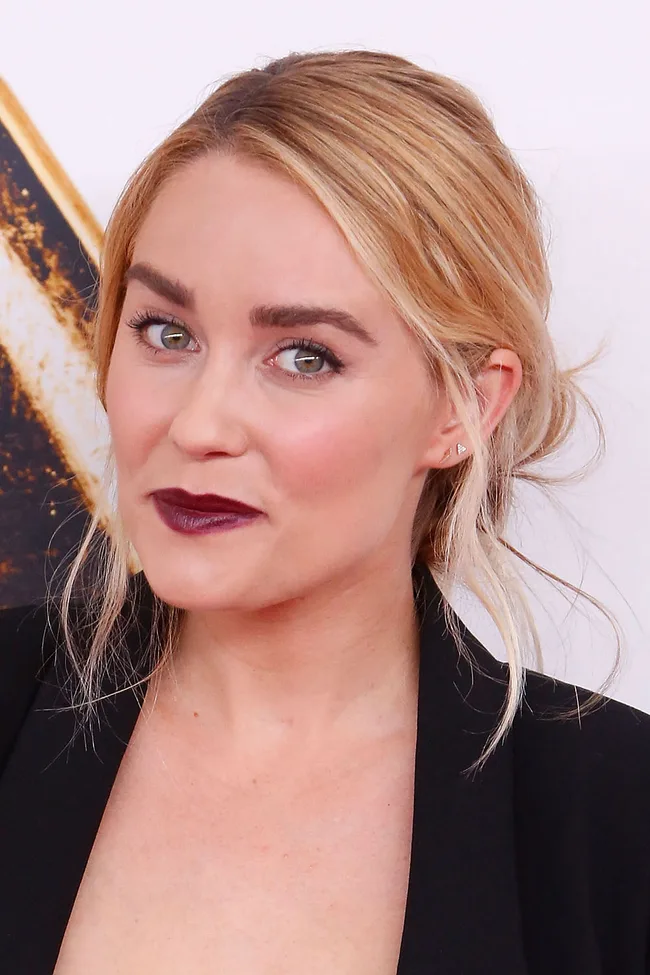 A person with blonde hair tied back, wearing dark lipstick and a black top, smiles slightly at the camera.