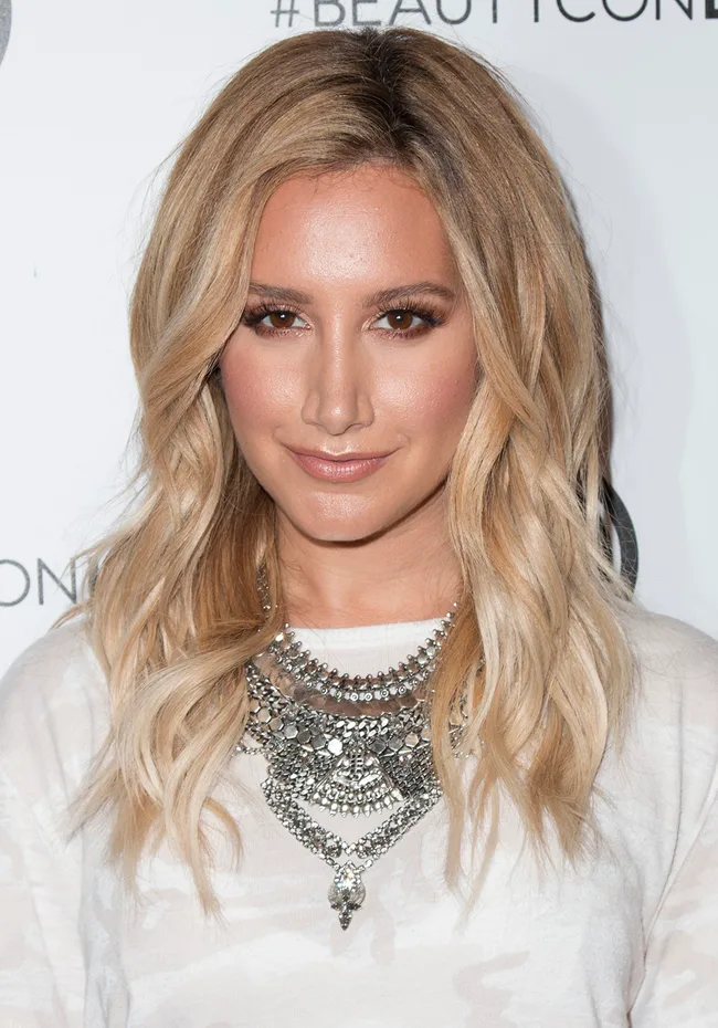 Woman with long blonde hair and layered necklace at an event, smiling softly at the camera.